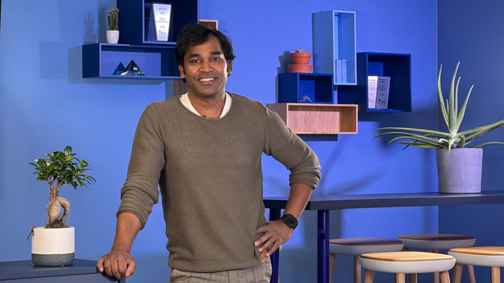 Ein Mann lächelt in einem blau eingerichteten Raum mit Pflanzen und Regalen.