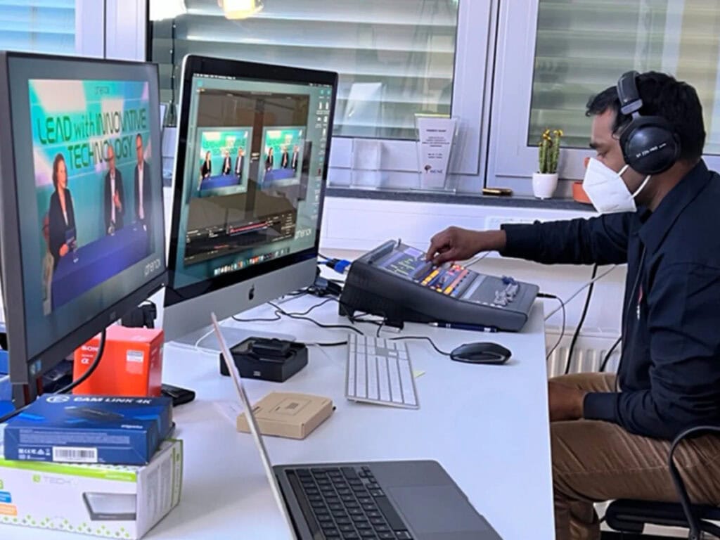 Mann mit Kopfhörern und Maske, bedient Mischpult; zwei Bildschirme zeigen Präsentationen.