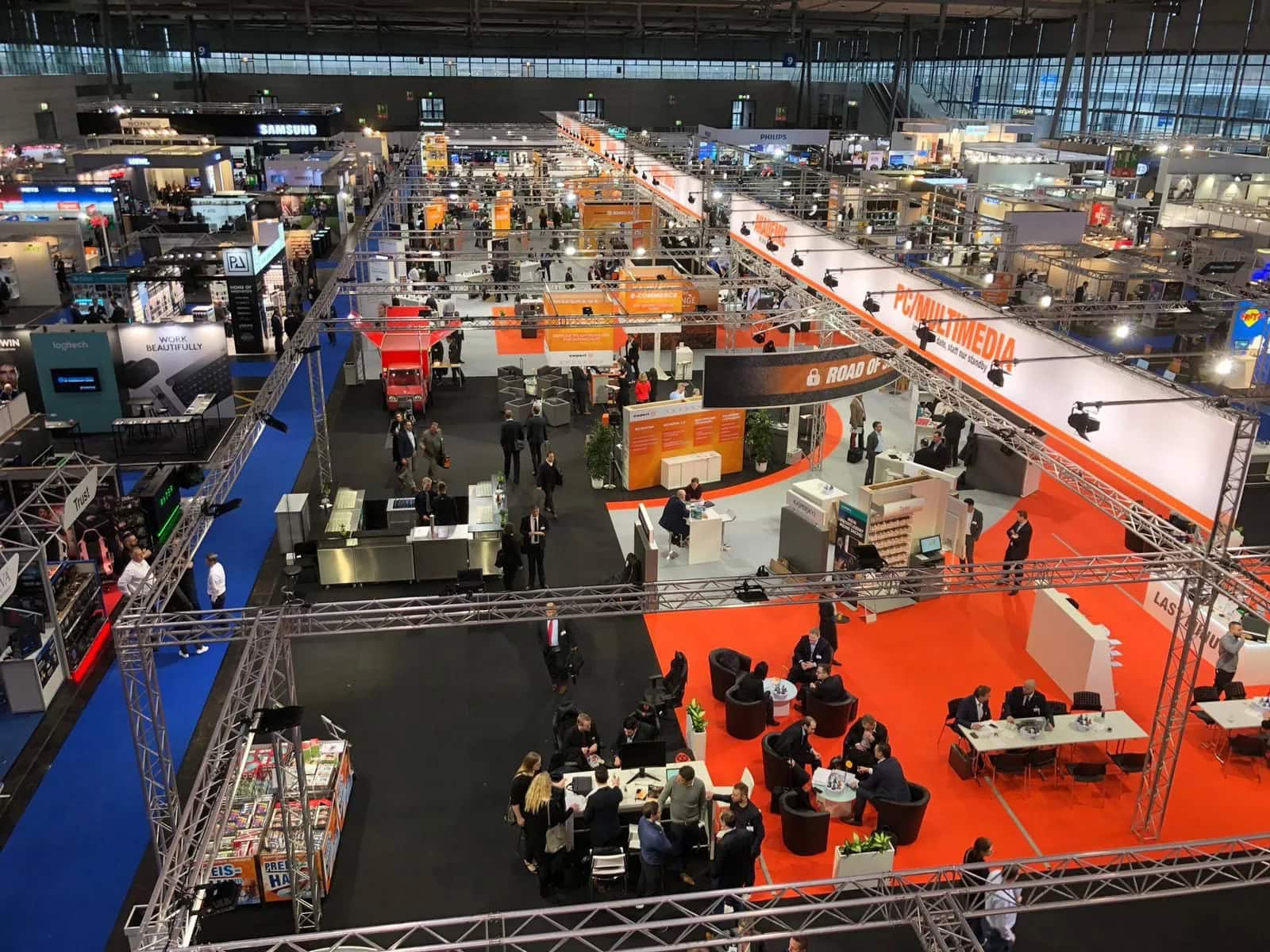 Vogelperspektive auf eine geschäftige Messehalle mit Ständen in leuchtenden Farben und Besuchern.