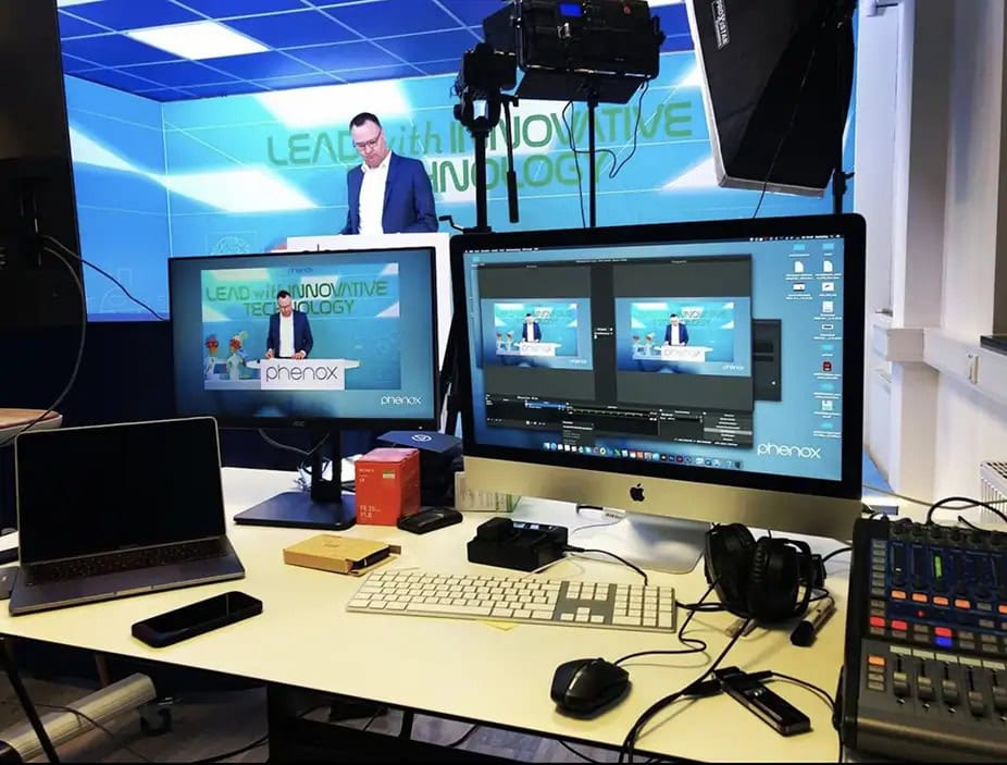 Ein Mann im Anzug präsentiert, lächelnd, vor blauer Technologie-Kulisse; Bildschirme im Vordergrund.